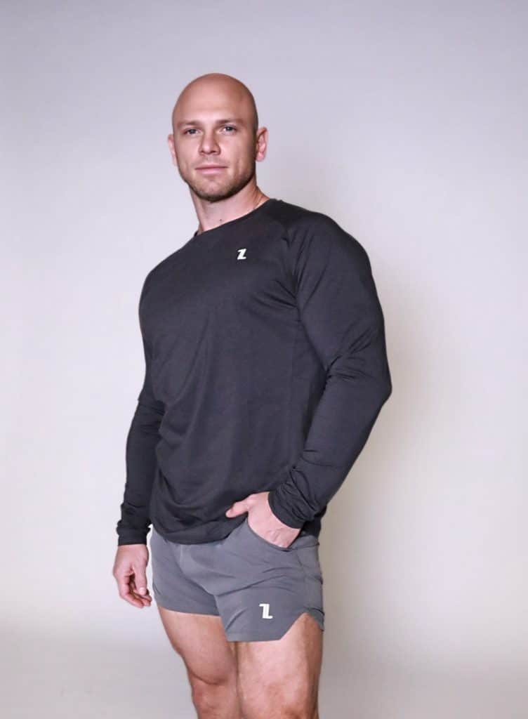 A muscular man with a shaved head wears a black long-sleeve shirt and gray athletic shorts, both with a small "Z" logo, standing confidently against a plain light background.