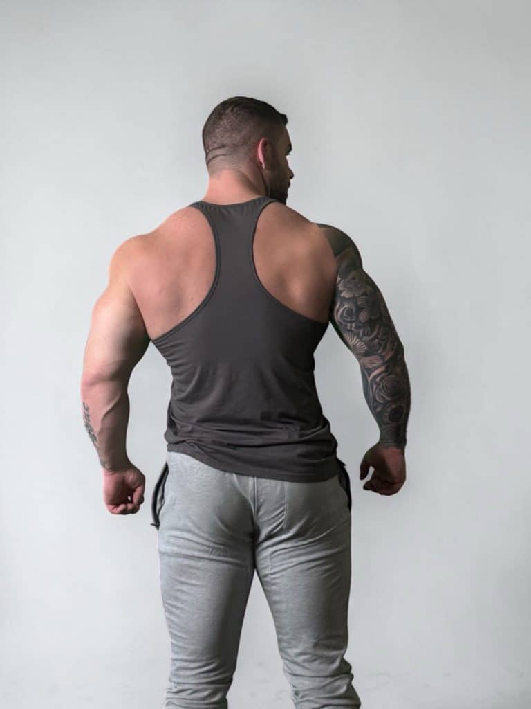 A muscular man facing away from the camera, wearing a dark tank top and light gray pants, showing a tattoo sleeve on his left arm against a plain light background.