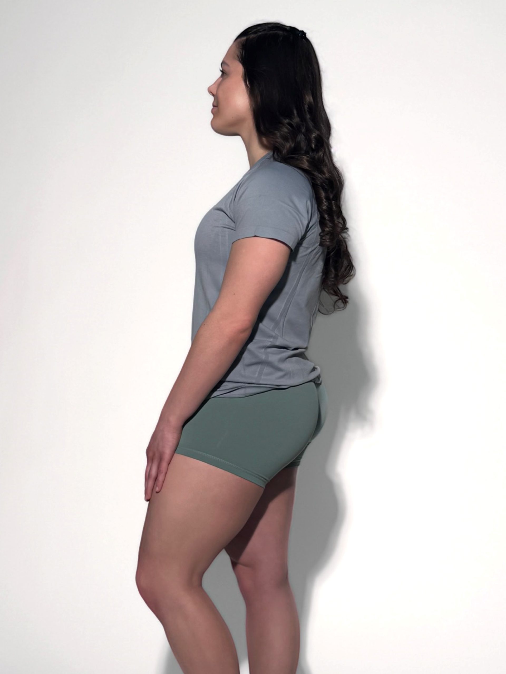 A woman stands in profile against a white background, wearing the GlideFlex Tee in gray and green shorts. Her long dark hair is down, and she poses with a relaxed posture, arms at her sides.