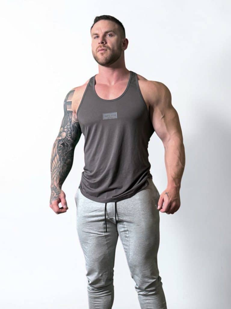 A muscular man wearing a gray tank top and light gray joggers stands confidently against a plain white background. One of his arms is heavily tattooed.