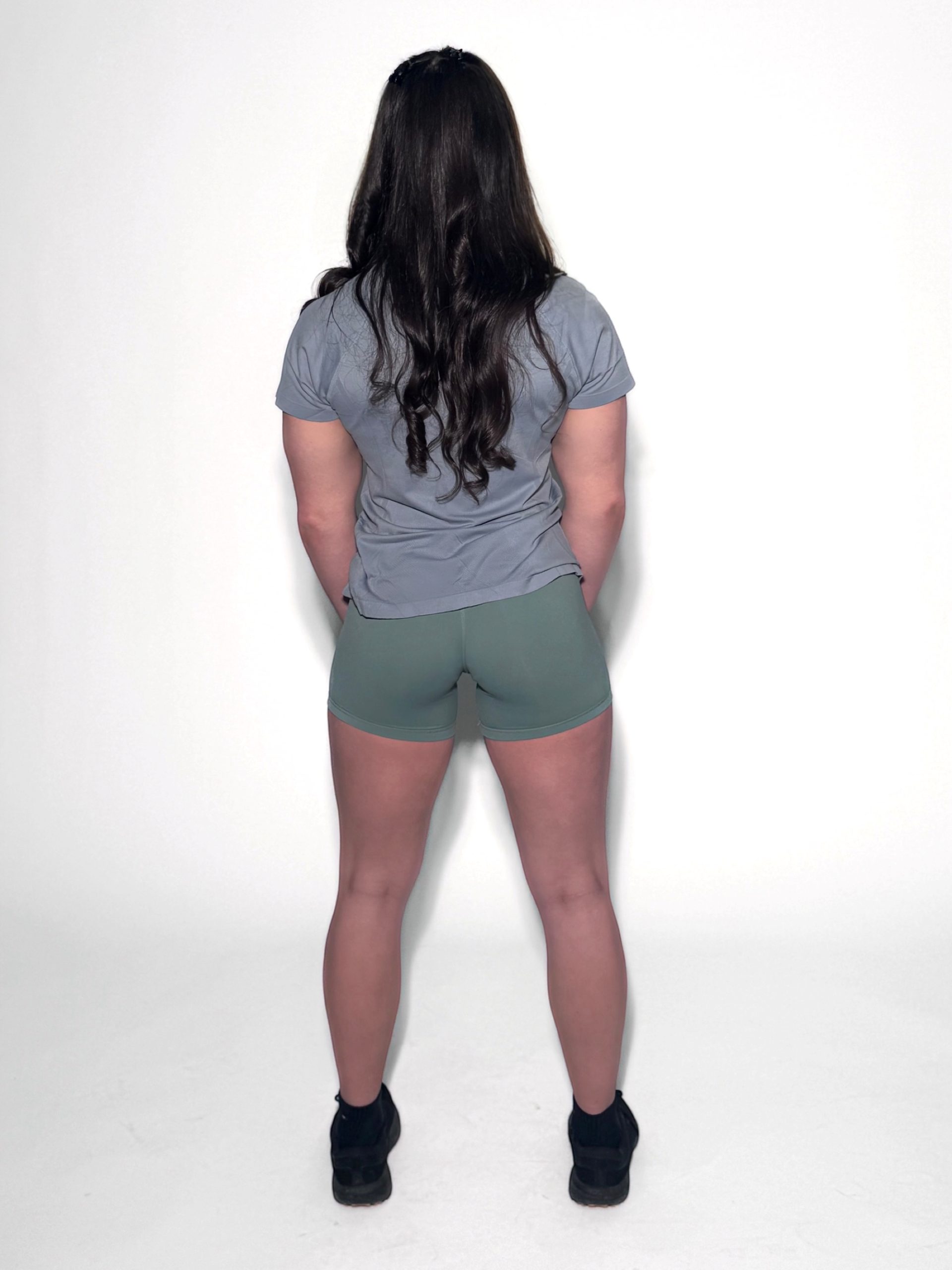 A person with long brown hair stands facing away from the camera, wearing a gray GlideFlex Tee, green shorts, and black shoes against a plain white background.