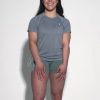 A woman with long dark hair smiles against a plain light background, wearing the gray GlideFlex Tee and green shorts, her hands resting on her thighs.