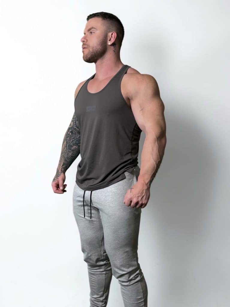 A muscular man with a beard, wearing a dark tank top and light gray joggers, stands against a plain white background, looking off to the side with his arms slightly flexed and hands relaxed.