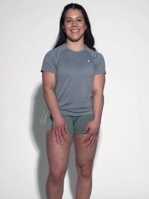 A woman with long dark hair smiles against a plain light background, wearing the gray GlideFlex Tee and green shorts, her hands resting on her thighs.