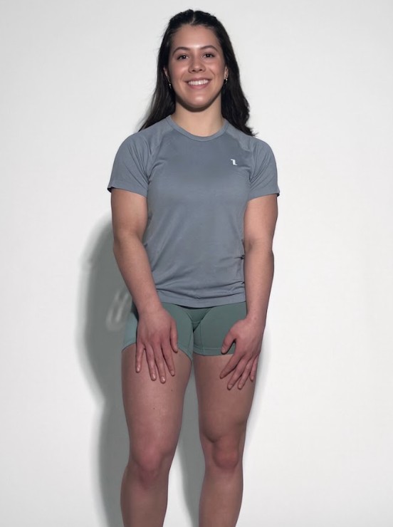 A woman with long dark hair smiles against a plain light background, wearing the gray GlideFlex Tee and green shorts, her hands resting on her thighs.