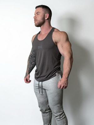 A muscular man with a beard and tattoos on his left arm stands against a plain white background, wearing a dark tank top and light gray sweatpants. He looks to the side with a relaxed expression.
