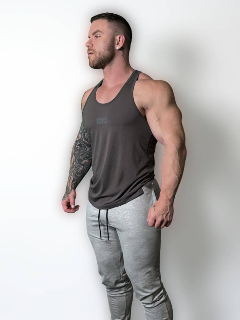 A muscular man with a beard and tattoos on his left arm stands against a plain white background, wearing a dark tank top and light gray sweatpants. He looks to the side with a relaxed expression.