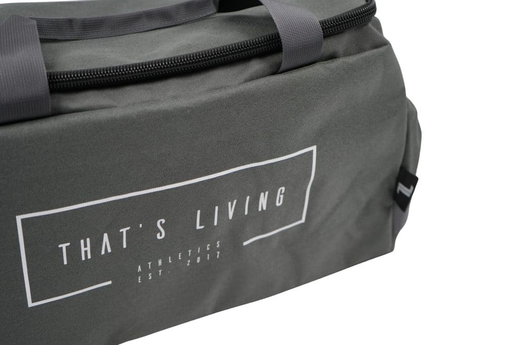 A close-up of the TRIARC Duffle Bag in gray, featuring black zippers and handles. The front displays printed white text: "THAT'S LIVING ATHLETICS EST. 2017.