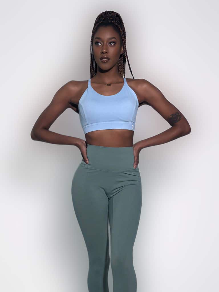 A woman models the Everyday Ease Bra in light blue and high-waisted sage green leggings, standing confidently against a plain light backdrop with her braided hair in a high ponytail and hands on her hips.