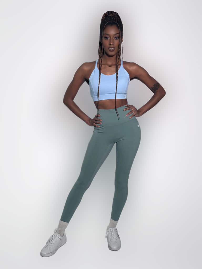 A woman stands confidently with hands on hips, wearing the Everyday Ease Bra in light blue, green leggings, white sneakers, and long braided hair, posed against a plain white background.