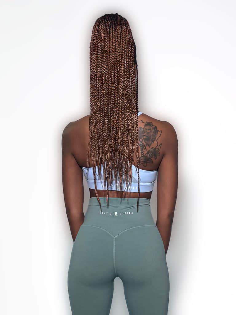 A woman with long braids, wearing the Everyday Ease Bra in light blue and green leggings, stands facing a plain white wall, revealing a large floral tattoo on her upper left back.