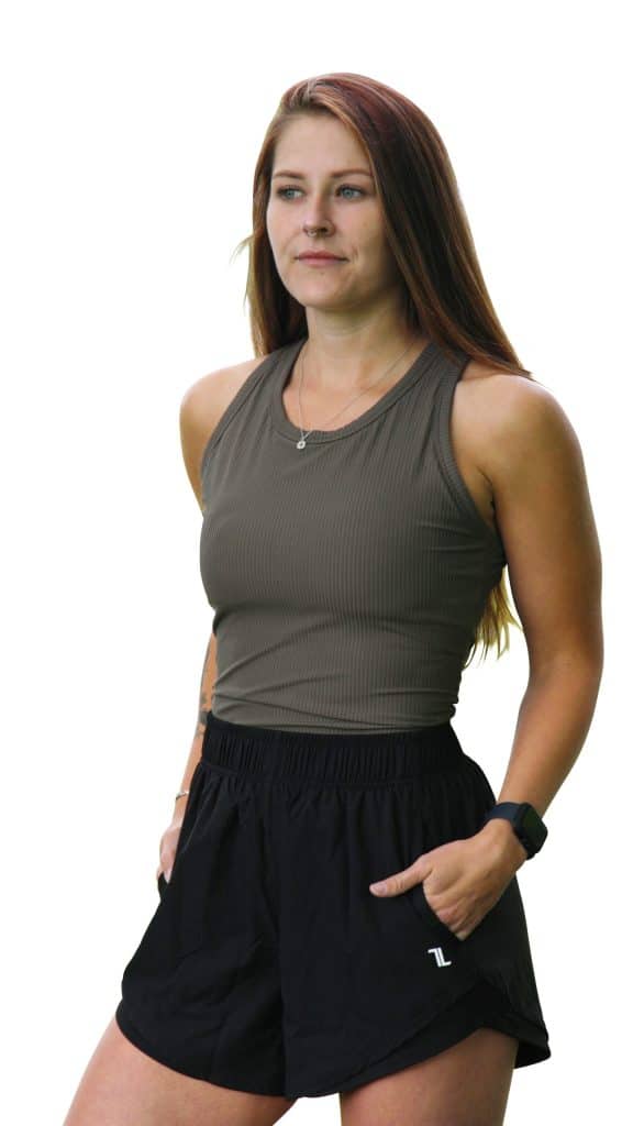 A woman with long brown hair stands with her hands in the pockets of her black athletic shorts, wearing a sleeveless ribbed tank top and a smartwatch, looking slightly to the side with a neutral expression.