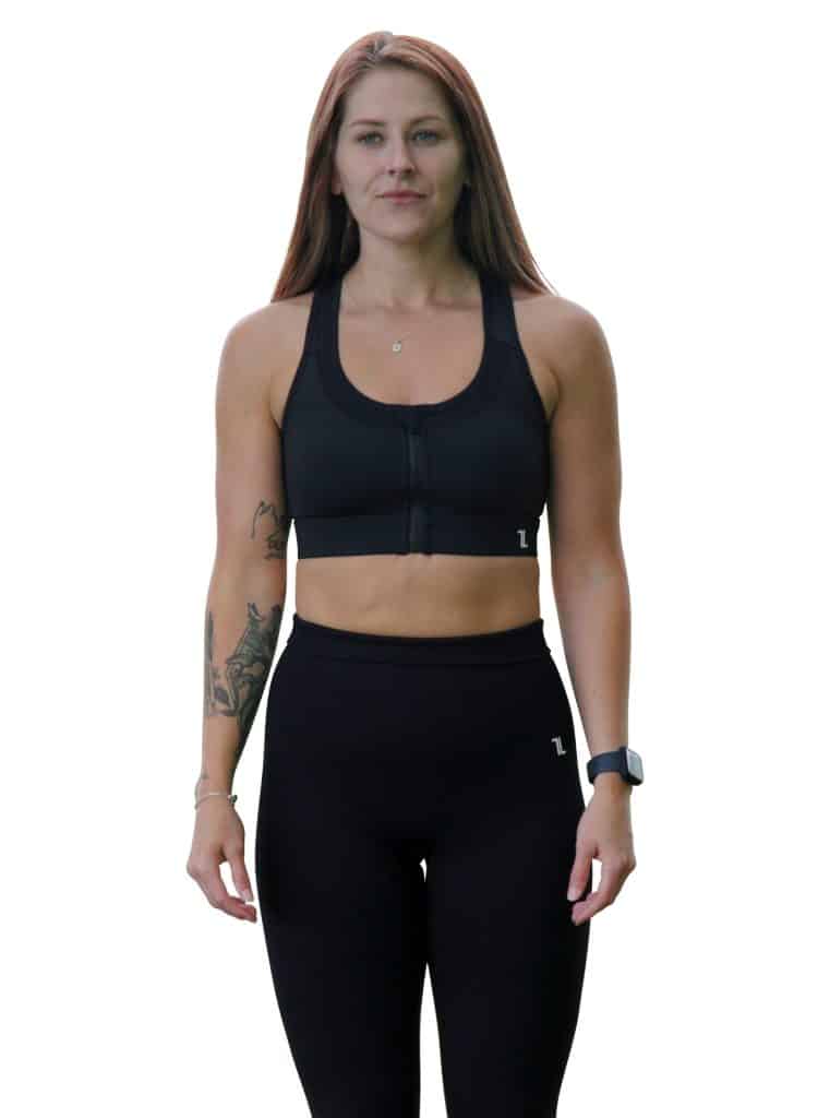 A woman with long brown hair wearing a black sports bra and black leggings stands facing forward. She has a visible tattoo on her left arm and is wearing a watch on her right wrist. The background is white.