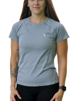 A woman with long brown hair wearing a light gray short-sleeve athletic shirt and black leggings stands facing forward. She has a tattoo on her left arm and is wearing a smart watch.