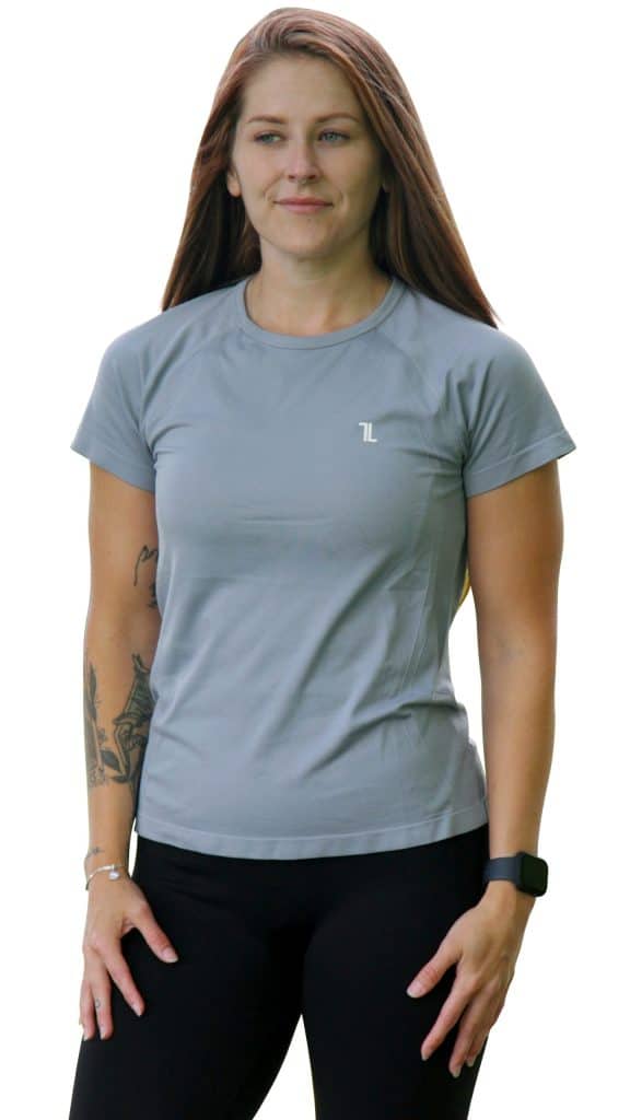 A woman with long brown hair wearing a light gray short-sleeve athletic shirt and black leggings stands facing forward. She has a tattoo on her left arm and is wearing a smart watch.