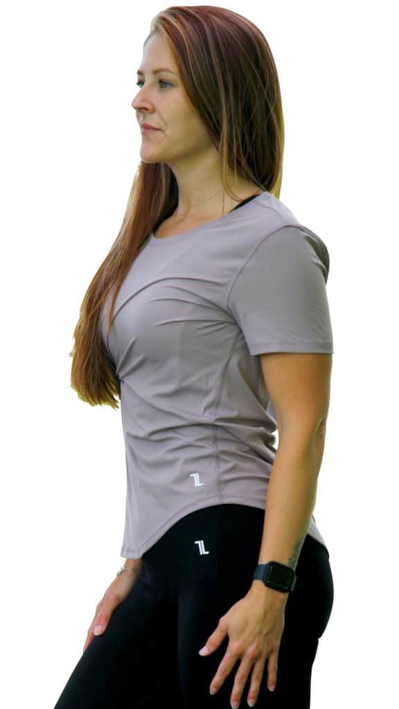 A woman with long brown hair stands sideways against a white background, wearing the VeloraFlow Tee in light gray with black leggings and a smartwatch. She looks slightly ahead with a neutral expression.