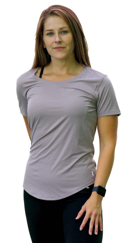 A woman with long brown hair wearing a light purple short-sleeve shirt, black leggings, and a smartwatch stands facing forward with a neutral expression against a white background.