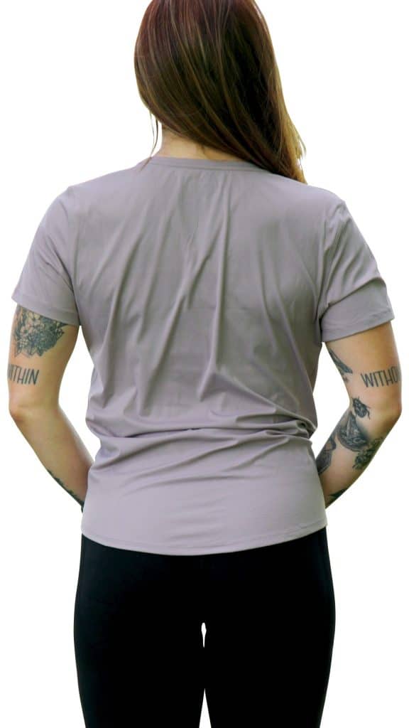 A person with long brown hair stands facing away, wearing the VeloraFlow Tee in light gray and black pants. Both arms feature visible tattoos against a white background.