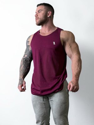 A muscular man with tattoos on his left arm models the Genesis Tank V2 in maroon and light gray sweatpants, standing against a white background and looking to the side.