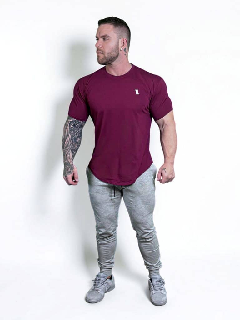 A muscular man with tattoos on his left arm stands against a white background, wearing a fitted maroon t-shirt, light gray jogger pants, and gray sneakers. He is looking to his left with his arms at his sides.