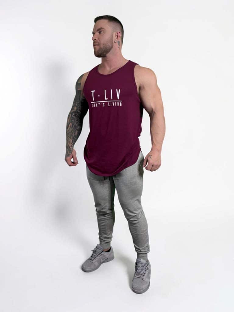 A muscular man wearing a maroon sleeveless T-shirt with "T·LIV THAT'S LIVING" on the front, gray jogger pants, and gray sneakers stands against a plain white background.