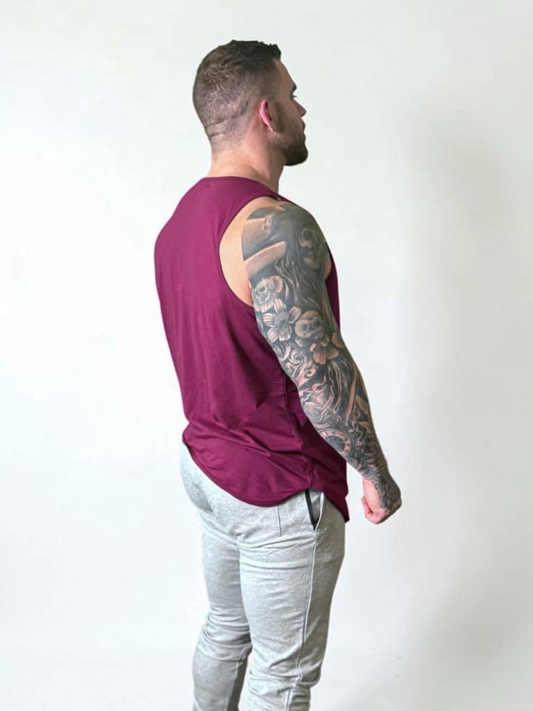 A man with short hair shows off his tattooed right arm while standing sideways in a plain white setting, wearing the sleeveless maroon Genesis Tank V2 and light gray pants.