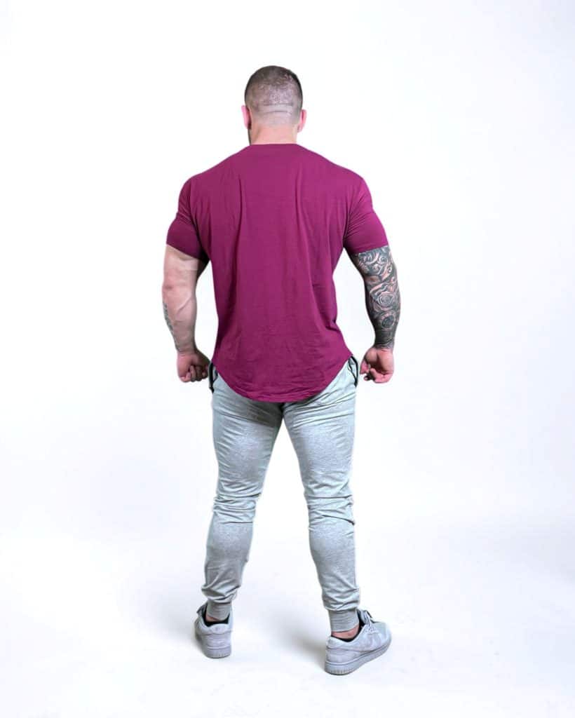 A muscular man stands facing away from the camera, wearing a maroon t-shirt, light gray jogger pants, and gray sneakers. His right arm has a large tattoo sleeve. The background is plain white.