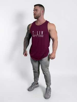 A muscular man with tattoos stands on a white background, wearing a burgundy sleeveless "T. LIV THAT'S LIVING" tank top, gray jogger pants, and gray sneakers, looking off to his left.