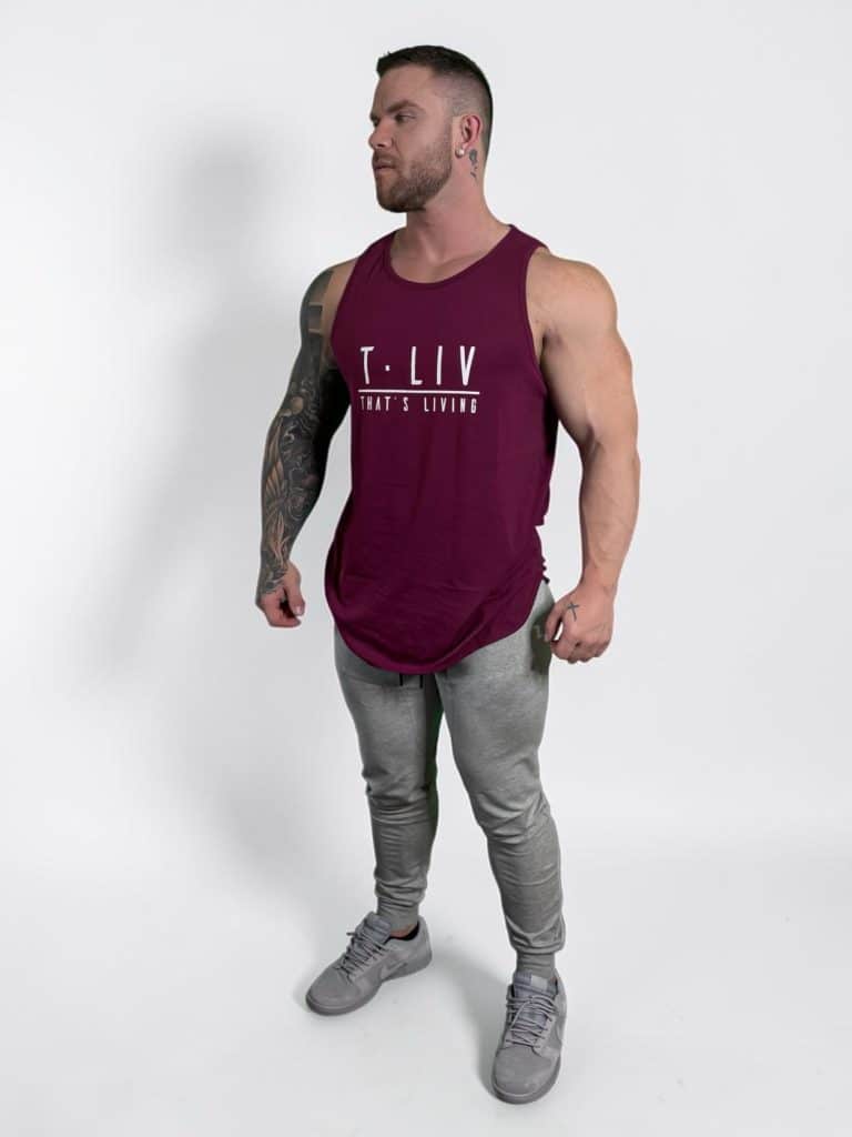 A muscular man with tattoos stands on a white background, wearing a burgundy sleeveless "T. LIV THAT'S LIVING" tank top, gray jogger pants, and gray sneakers, looking off to his left.