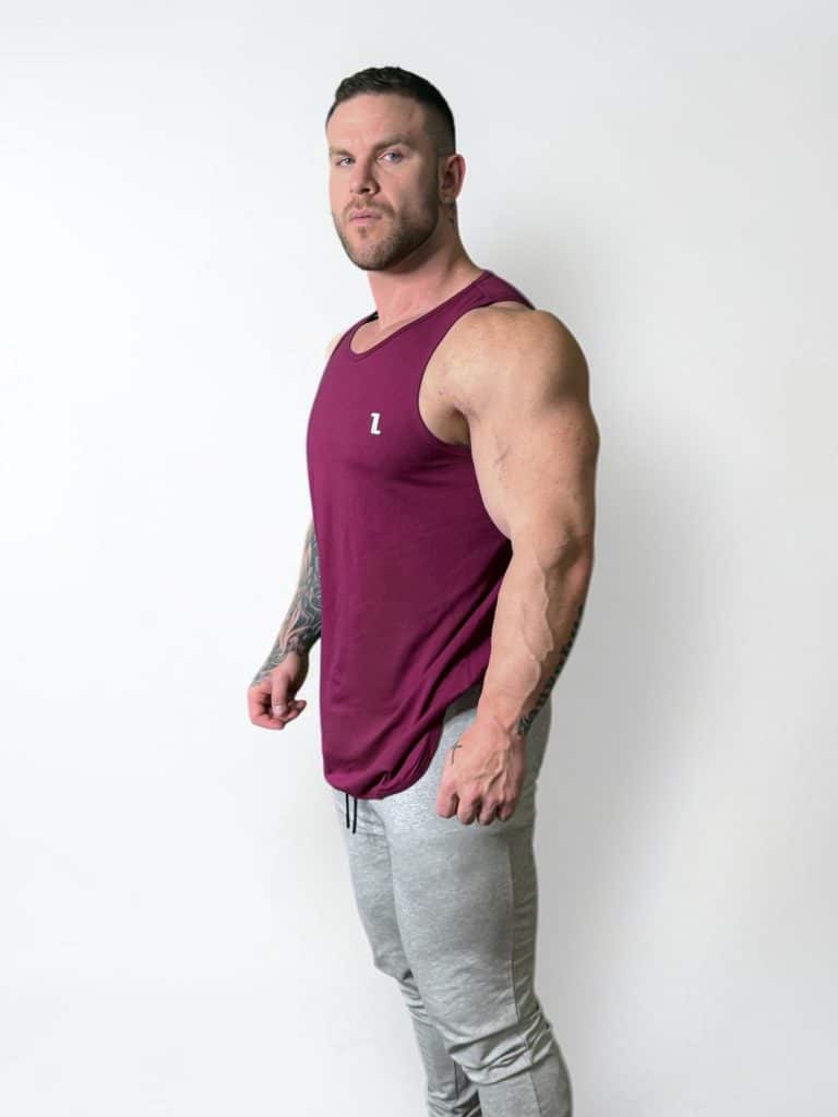 A muscular man with short hair models the maroon Genesis Tank V2 and light gray joggers, posing confidently against a plain white background.