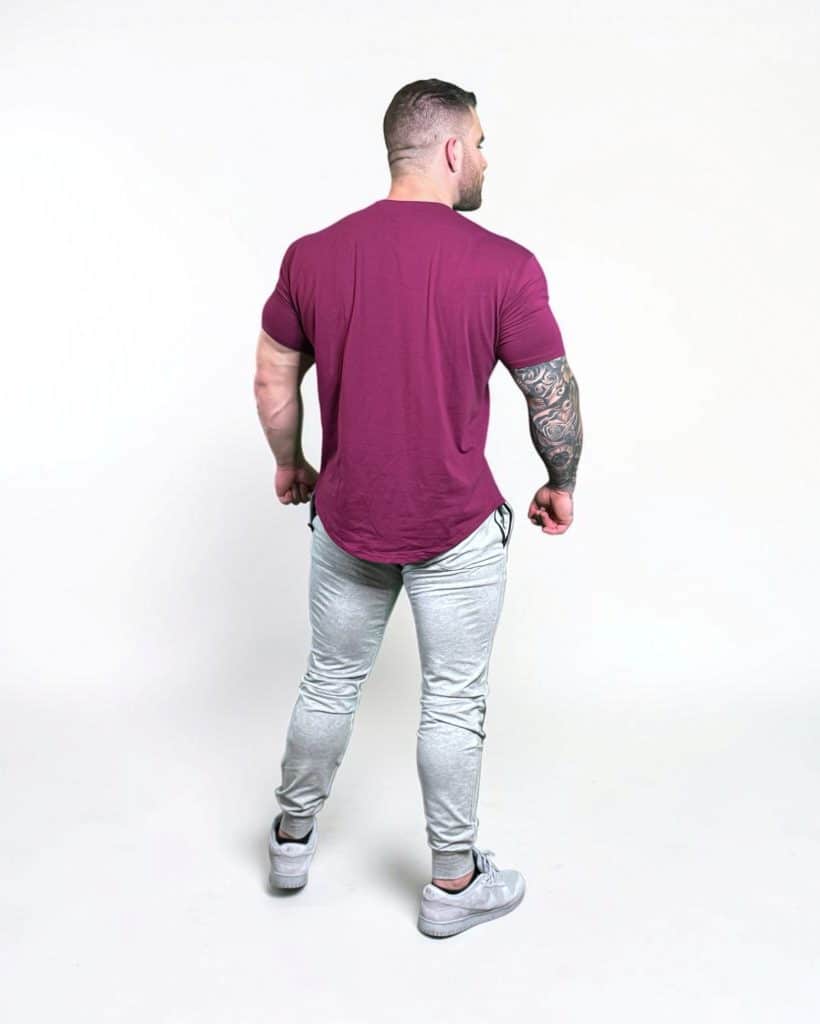 A man with short hair stands facing away, wearing a maroon T-shirt, light gray jogger pants, and gray sneakers. His right arm has a sleeve tattoo. The background is plain white.