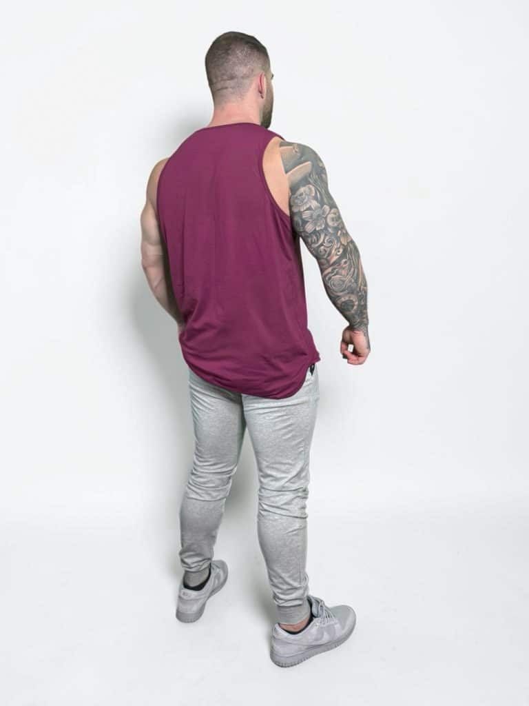 A muscular man with tattooed arms stands facing away, wearing a sleeveless maroon shirt, light gray joggers, and gray sneakers against a plain white background.