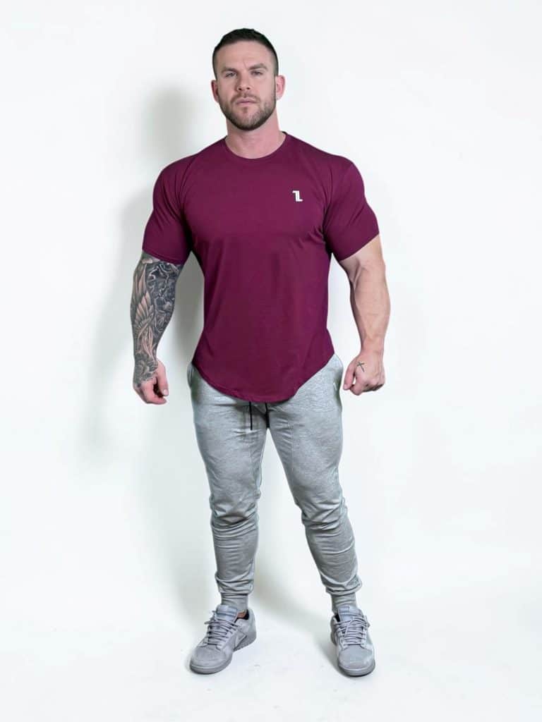 A muscular man stands against a plain white background, wearing a fitted maroon T-shirt, light gray jogger pants, and gray athletic shoes. His right arm displays a large black tattoo sleeve.
