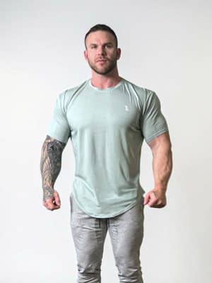 A muscular man with short hair and a beard stands against a plain white background, wearing a light gray fitted T-shirt and gray sweatpants. One of his arms features a detailed tattoo sleeve.