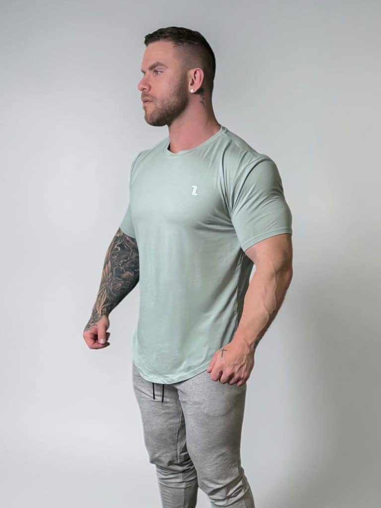 A muscular man with a full sleeve tattoo on his left arm stands against a plain light background, wearing a light green fitted t-shirt and gray jogger pants, looking off to the side with a serious expression.