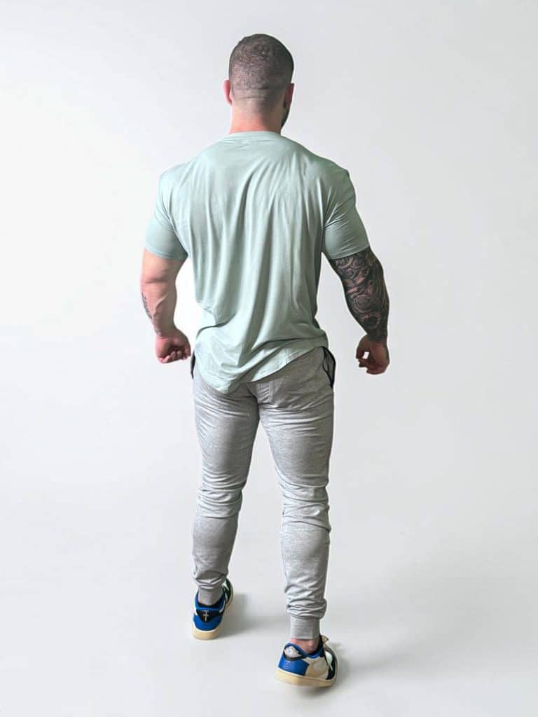 A muscular man with tattooed right arm stands facing away, wearing a light green t-shirt, gray joggers, and blue sneakers, against a plain white background.