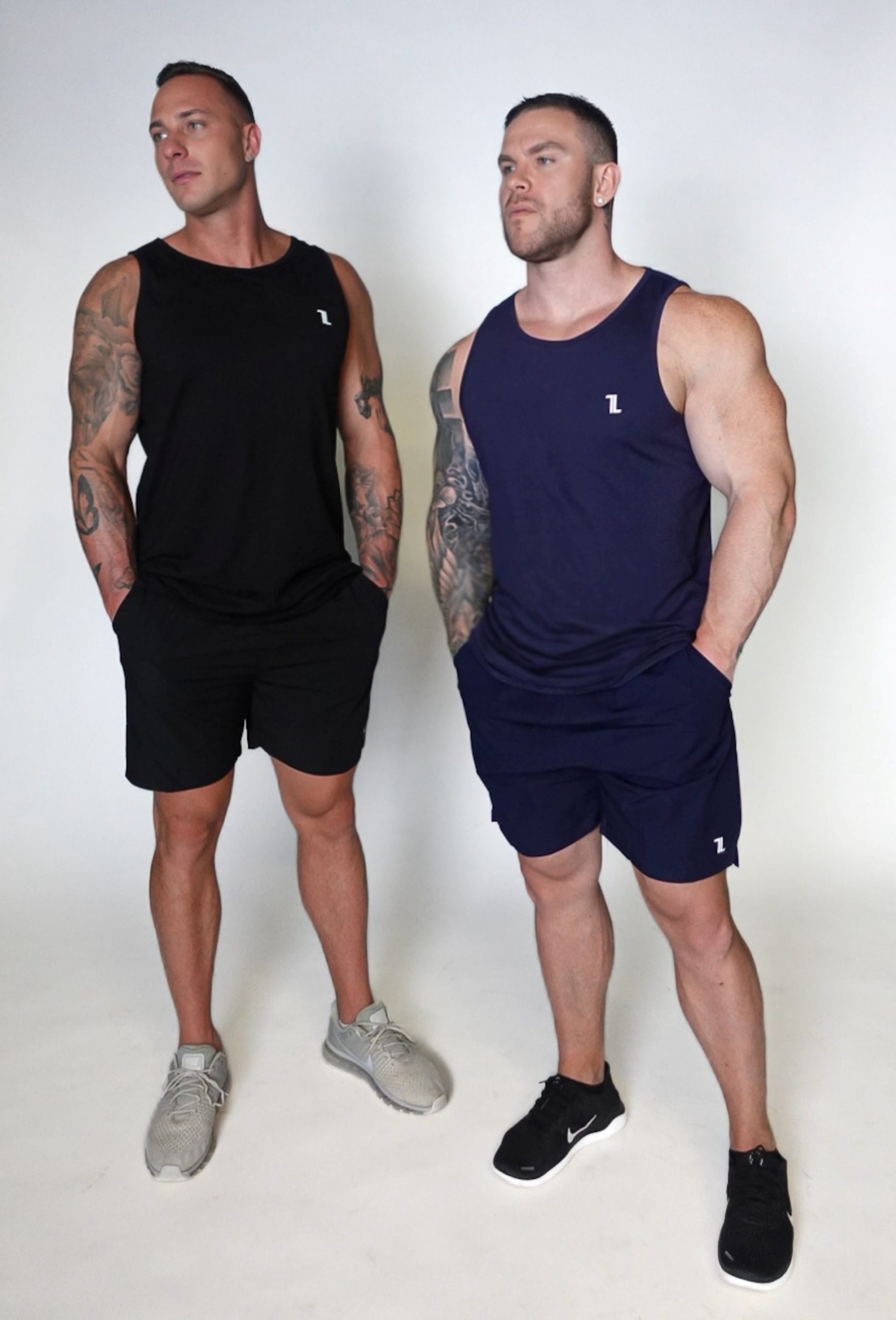 Two muscular men wear Genesis Tank V2 tops and shorts against a plain background. The left man wears black with grey sneakers; the right wears navy with black sneakers.