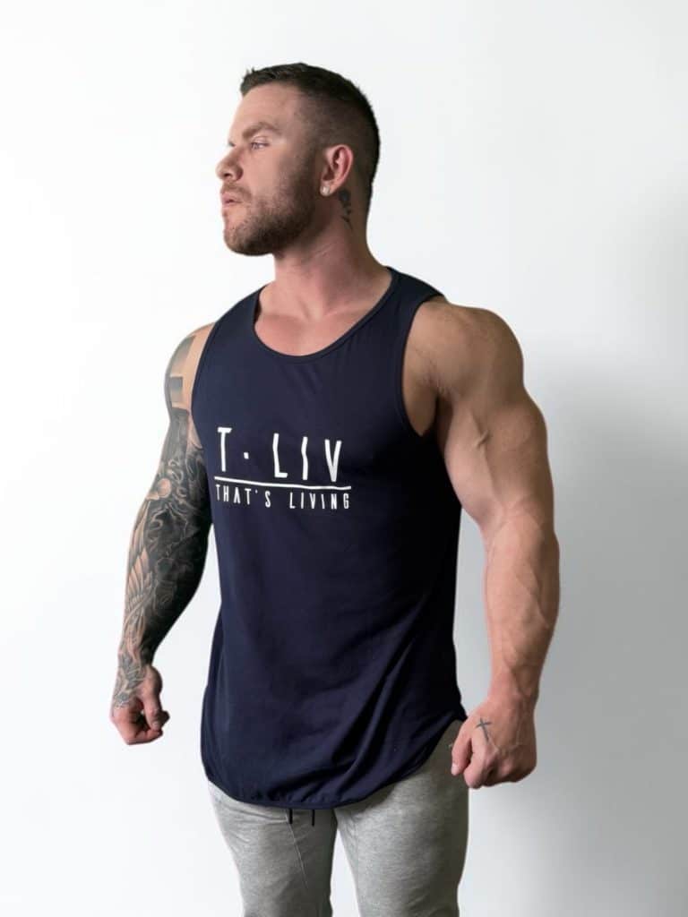 A muscular man with tattoos on his left arm wears the Genesis Tank, a navy top with "T. LIV THAT'S LIVING" on the front, and gray pants. He stands against a white background, looking left with clenched fists.