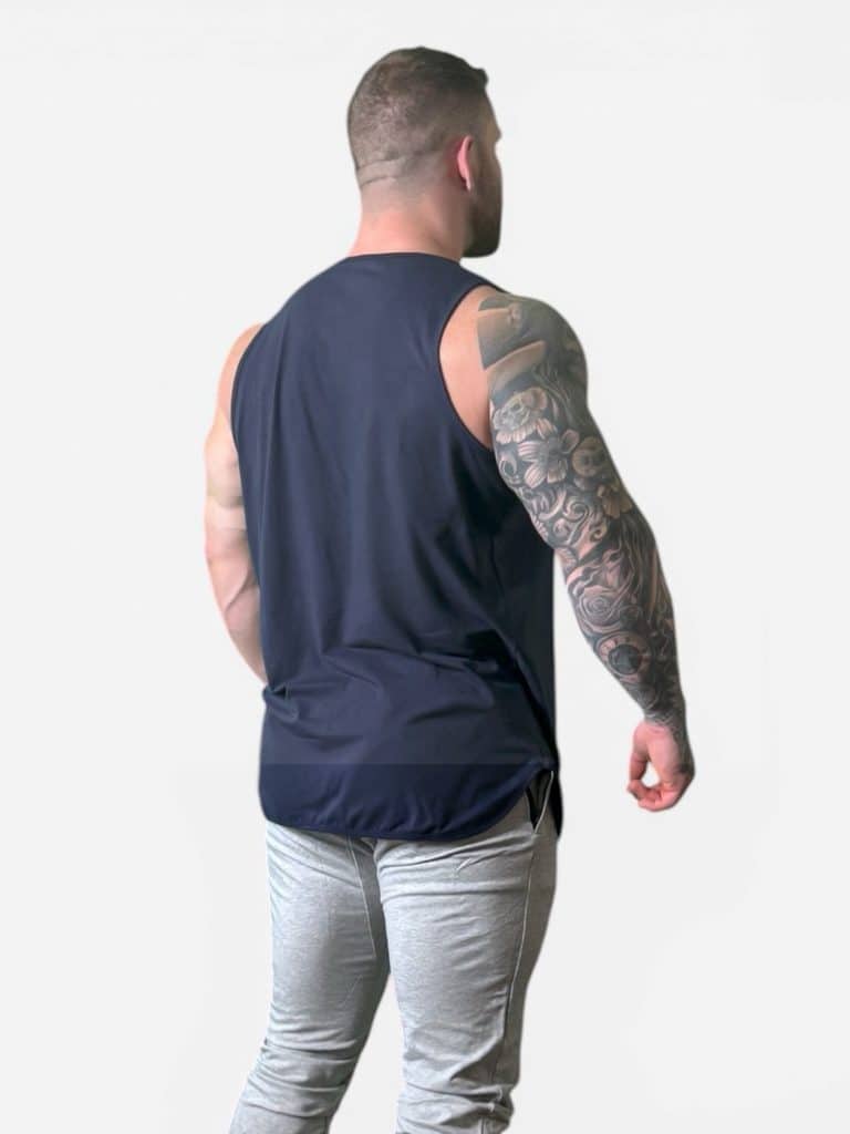 A muscular man with a tattooed right arm faces away, wearing the Genesis Tank in navy blue and light gray pants, standing against a plain light background.