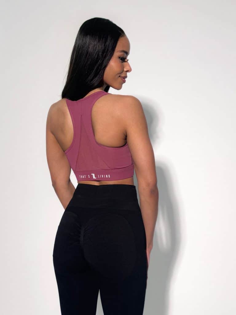 A woman stands facing away from the camera, wearing a mauve sports bra and black leggings, with long straight hair and her right hand resting by her side against a plain white background.