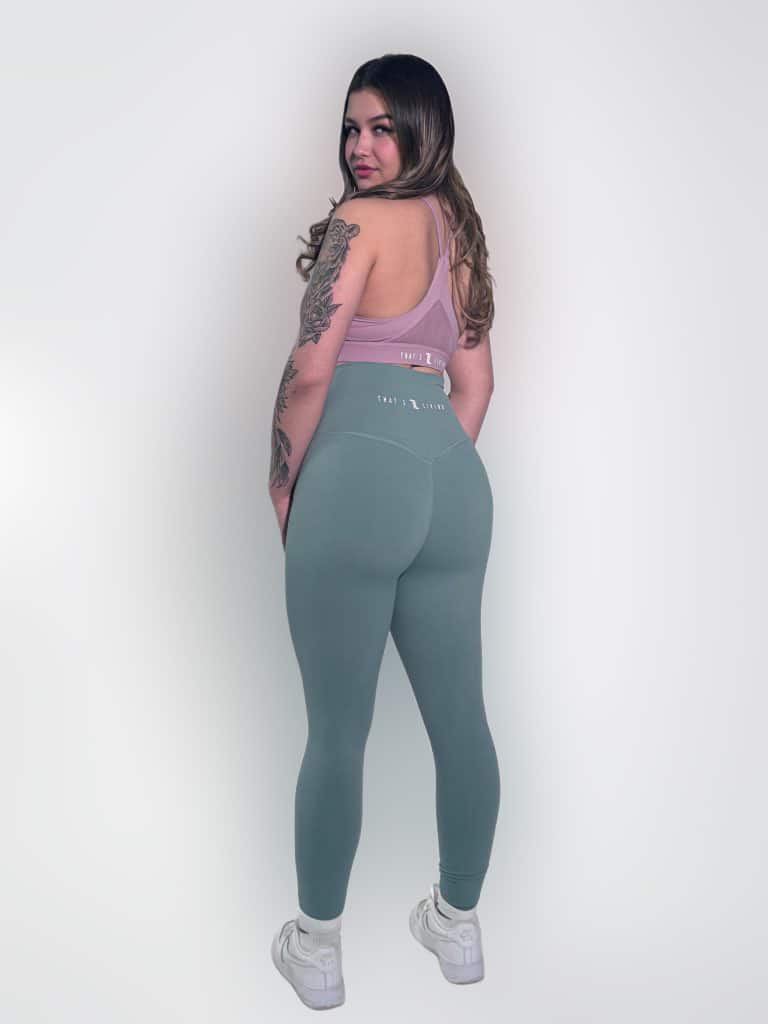 A woman with long hair and arm tattoos stands facing slightly backward, wearing a mauve sports bra, teal leggings, and white sneakers, against a plain light background.