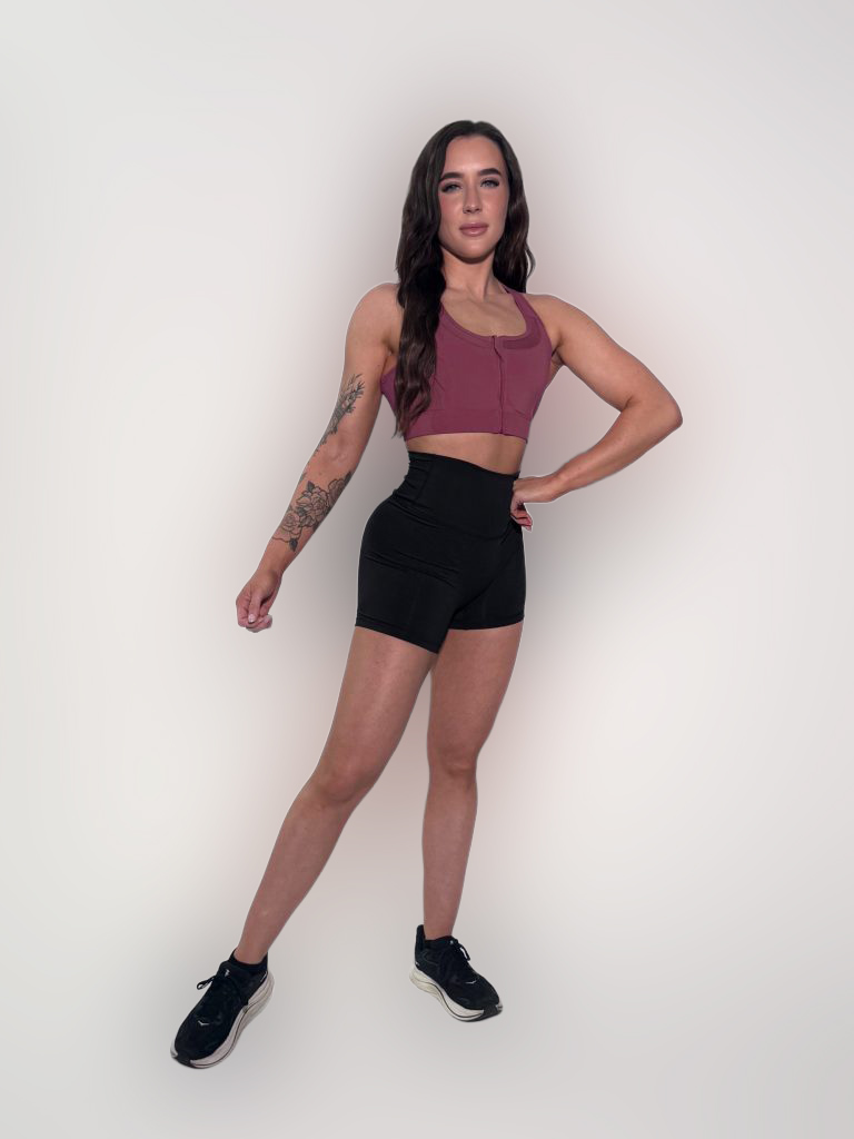A woman with long dark hair stands against a plain white background, wearing a mauve crop top, black high-waisted shorts, and black sneakers. She has a tattoo on her left forearm and is posing confidently with one hand on her hip.