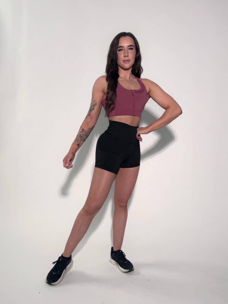 A woman with long dark hair stands against a plain white background, wearing a mauve crop top, black high-waisted shorts, and black sneakers. She has a tattoo on her left forearm and is posing confidently with one hand on her hip.