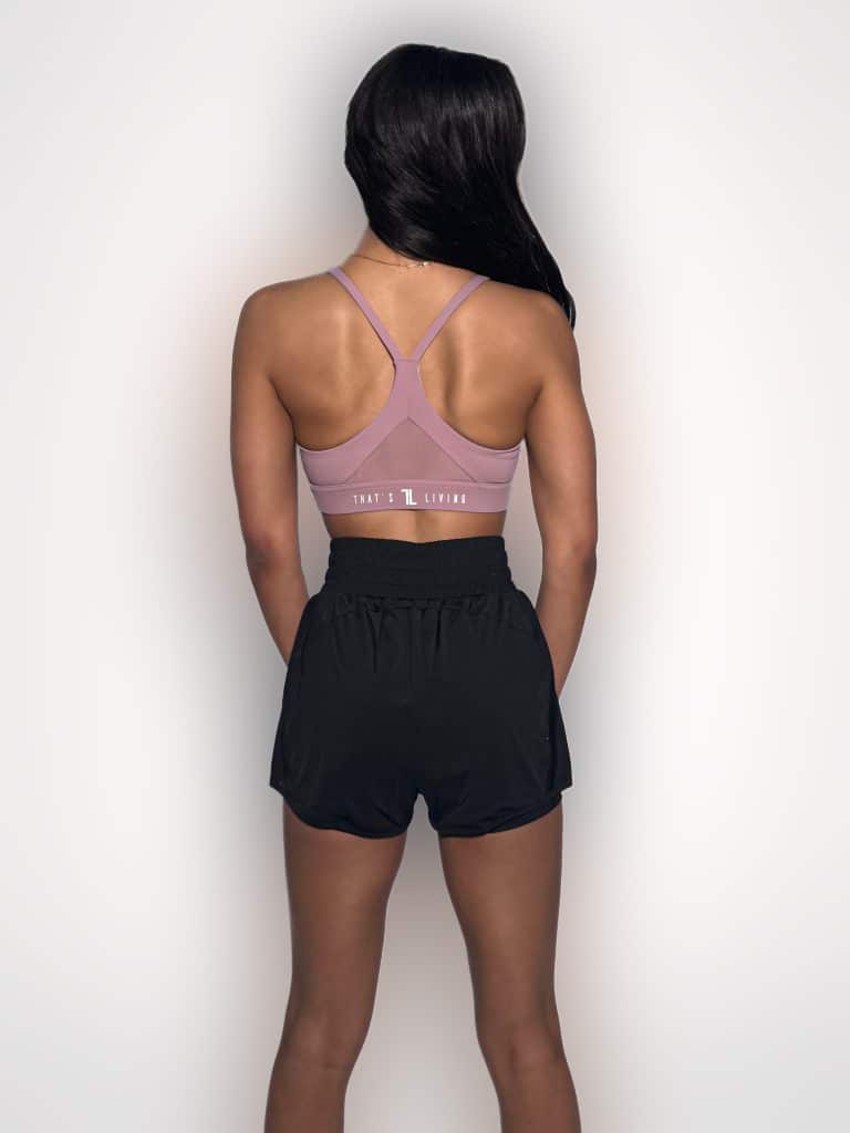A woman stands facing away from the camera, wearing a mauve sports bra and black athletic shorts. Her long dark hair is over one shoulder, and the background is plain white.