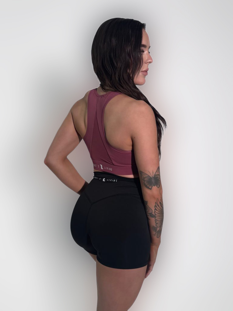 A woman with long dark hair stands sideways, looking left. She wears a maroon sports top and black athletic shorts. Her left arm shows visible tattoos of flowers and a butterfly. The background is plain white.