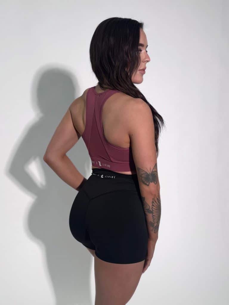 A woman with long dark hair stands sideways, looking left. She wears a maroon sports top and black athletic shorts. Her left arm shows visible tattoos of flowers and a butterfly. The background is plain white.