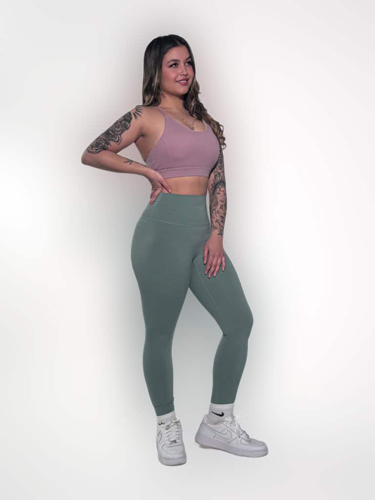 A woman with long hair and arm tattoos poses smiling in activewear—a mauve sports bra, high-waisted sage green leggings, and white sneakers—against a plain white background.