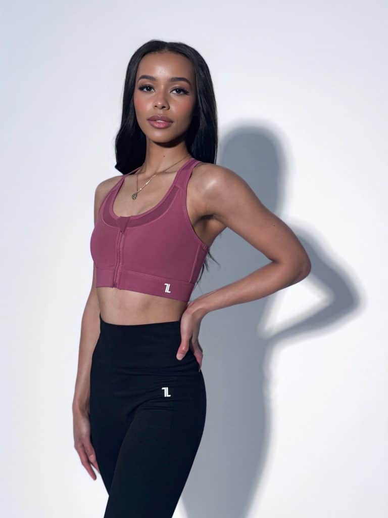 A woman stands confidently against a plain background, wearing a mauve sports bra and black leggings. Her long dark hair is down, and she has one hand on her hip, looking slightly to the side and smiling softly.