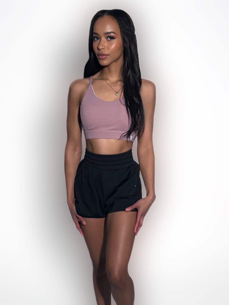 A woman stands against a plain white background, wearing a light purple crop top and black athletic shorts. She has long dark hair and poses with her hands resting on her thighs, looking confidently at the camera.