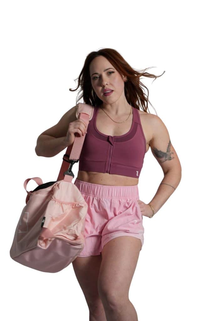 A woman with long brown hair, wearing a maroon sports bra and pink shorts, confidently poses against a white background with the TRIARC Duffle Bag in light pink on her shoulder.